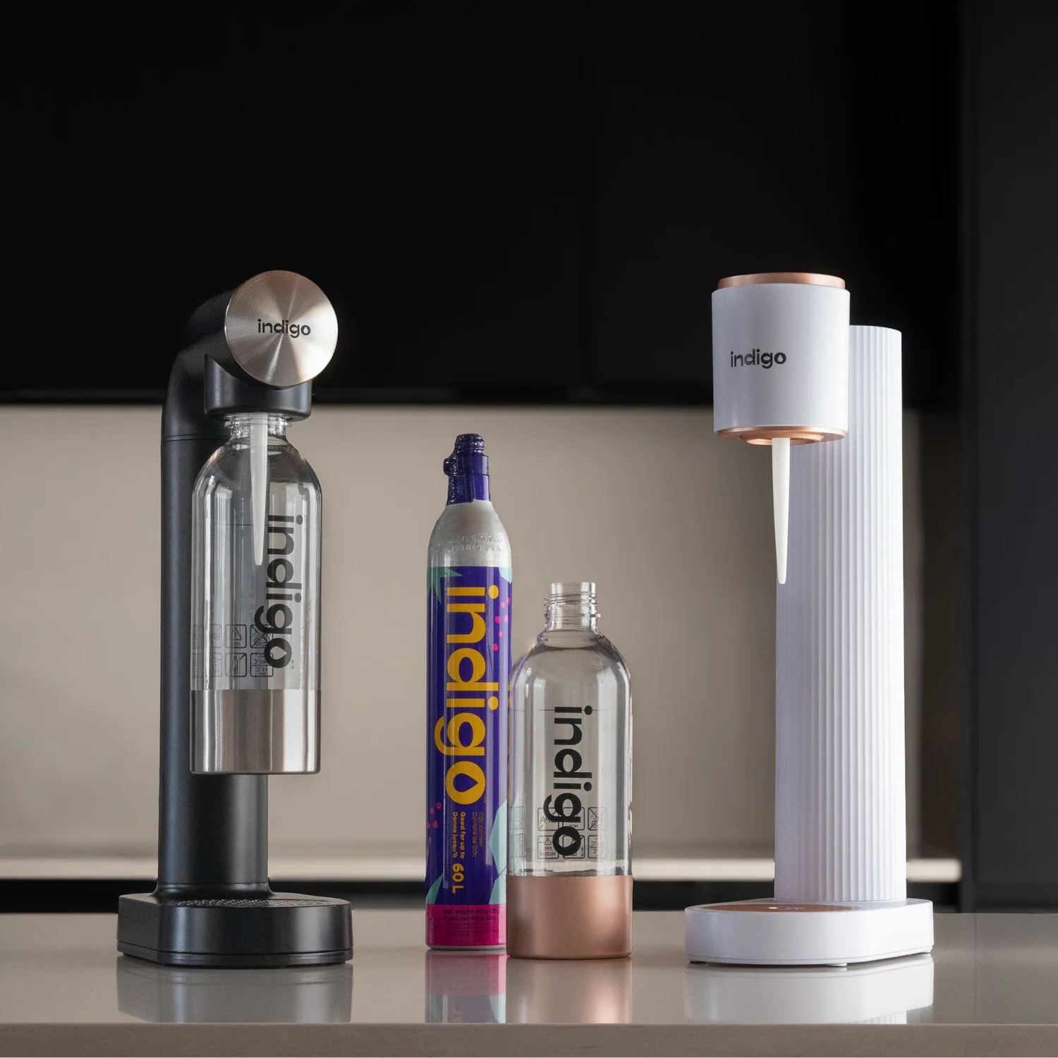 Machines à eau pétillante d'Indigo Soda : un modèle noir avec dessus en acier inoxydable et un modèle blanc à accents or rose, présentés avec une bouteille de CO2 et des bouteilles réutilisables sur un comptoir de cuisine.