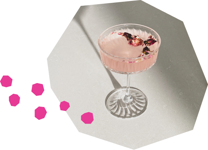 A pale pink sparkling water cocktail in a textured glass bowl, decorated with dried rose petals. The drink rests on a white surface, with a drop shadow creating a play of light.