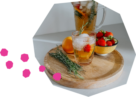 Un verre à cocktail décoré d'un brin de romarin, rempli d'un thé glacé pétillant et garni de fraises et d'une tranche d'orange. À côté du verre, sur un plateau en bois, se trouvent une orange entière, un bol rempli de fraises et un brin de romarin. En arrière-plan, on aperçoit un pichet rempli de la même boisson ambrée.